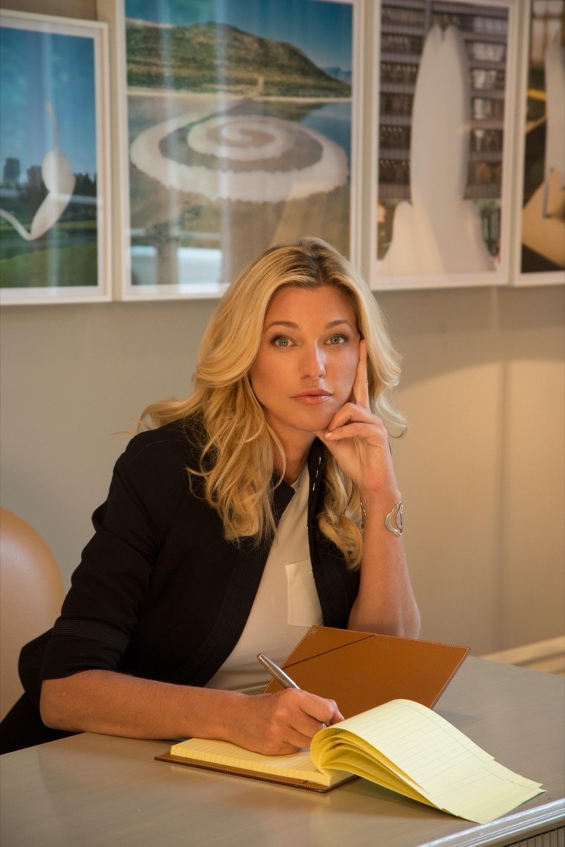 Nicole Junkerman in a black blazer, writing in a notebook at her desk with framed prints behind her